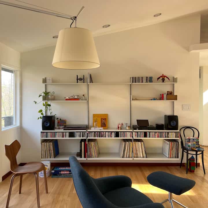 A white and brown toned living room featuring music speakers, bookshelves, and comfortable chairs in a minimal mid-century modern style