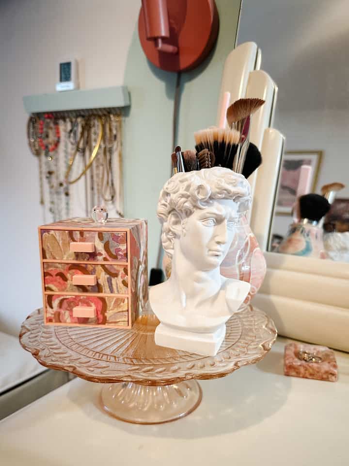 Pastel and white toned vanity featuring a vintage glass cake pedestal and sculptural makeup brush holder for an Instagrammable storage display