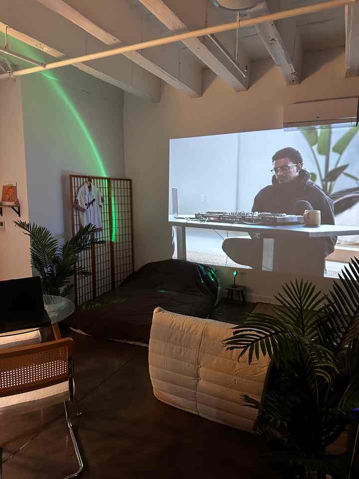 A wood-toned, minimal living room featuring a projector screen, rattan chair, and lazy floor sofa creating a cozy Japandi studio vibe