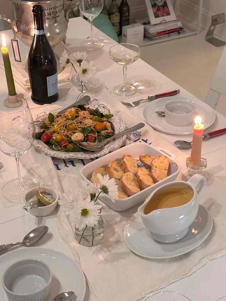 White tablecloth dining table featuring plates, glasses, salad pasta with seafood, bread, and lit candles creating cozy home party atmosphere