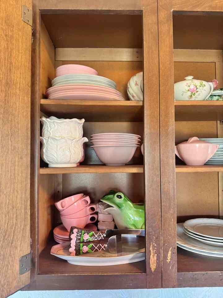 A cozy vintage kitchen cabinet featuring pink ceramic plates and a distinct frog pitcher inside wooden shelves