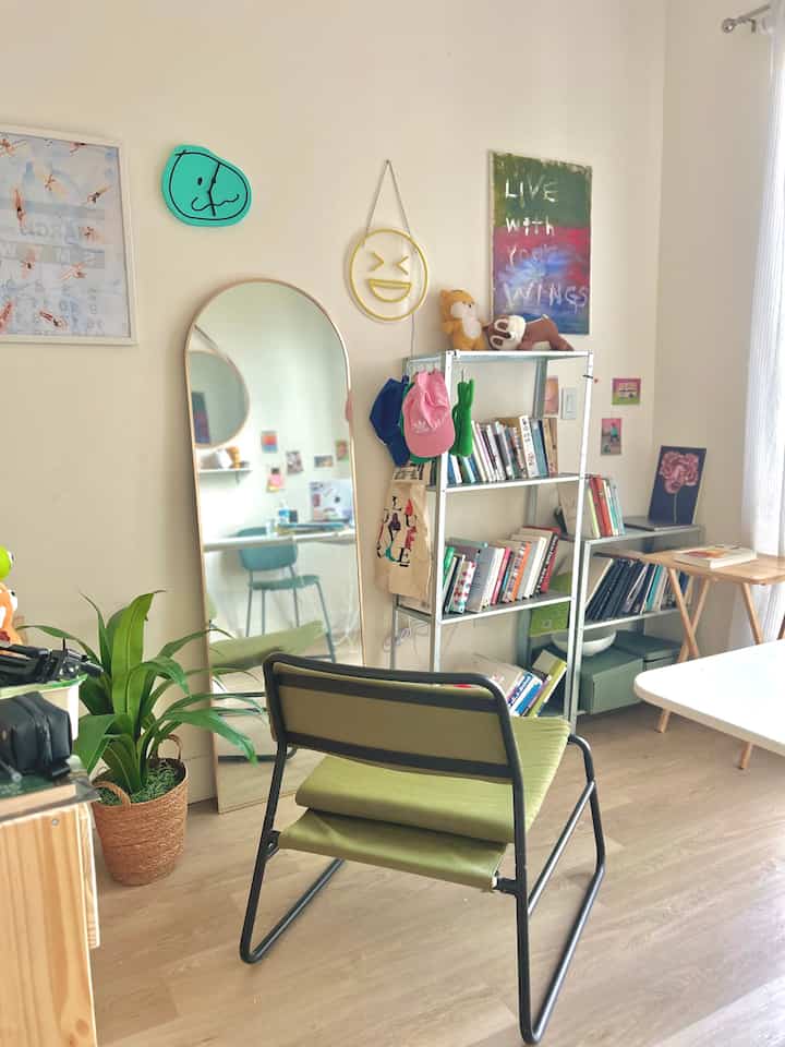 Natural-toned single household home office with beige walls, wooden floor, green chair, metal shelving unit, and arched full-length mirror creating cozy atmosphere