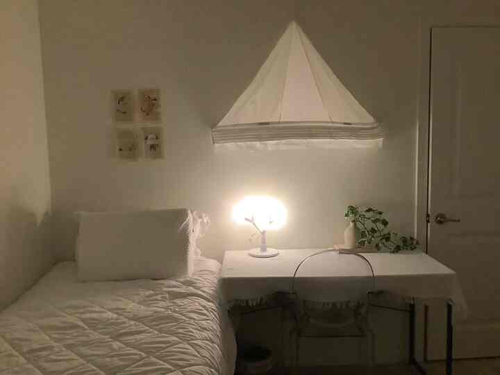 A white-toned bedroom featuring a simple bed and desk with a softly glowing table lamp creating a calm cozy atmosphere