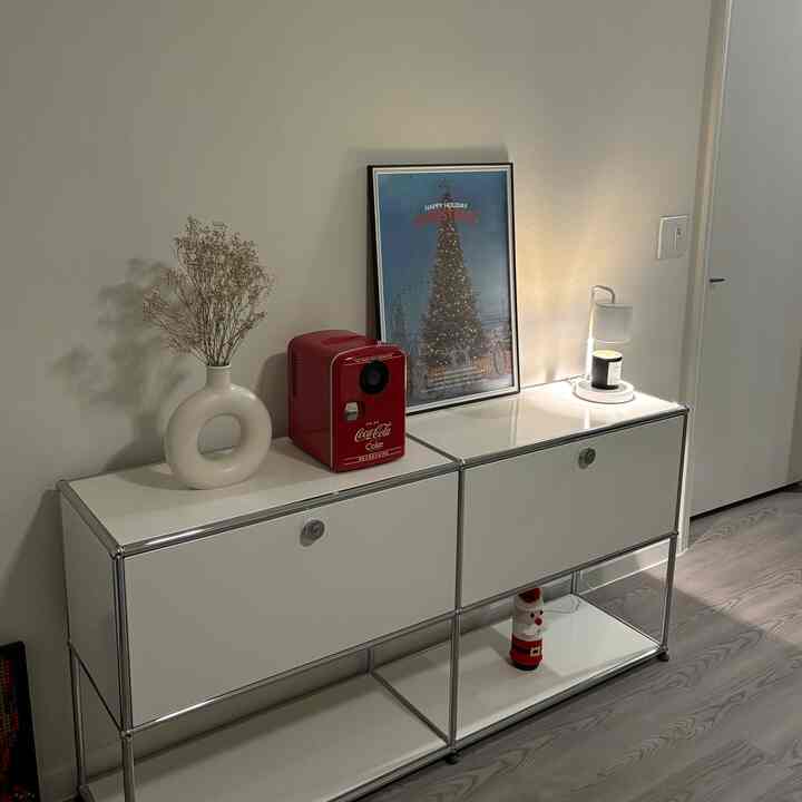 White-toned room with simple sideboard featuring red mini fridge, vase, poster, and candle in a minimal interior setting