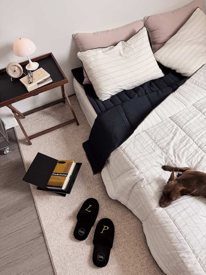 Beige and white toned bedroom featuring a bed, nightstand with table lamp and clock, with a dog resting comfortably