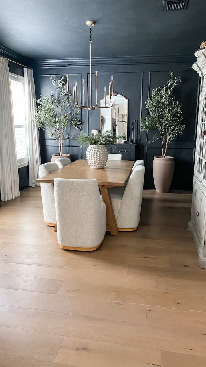Dark gray toned dining room featuring a wood tone dining table with white upholstered chairs, gold chandelier, and large plants creating a sophisticated atmosphere