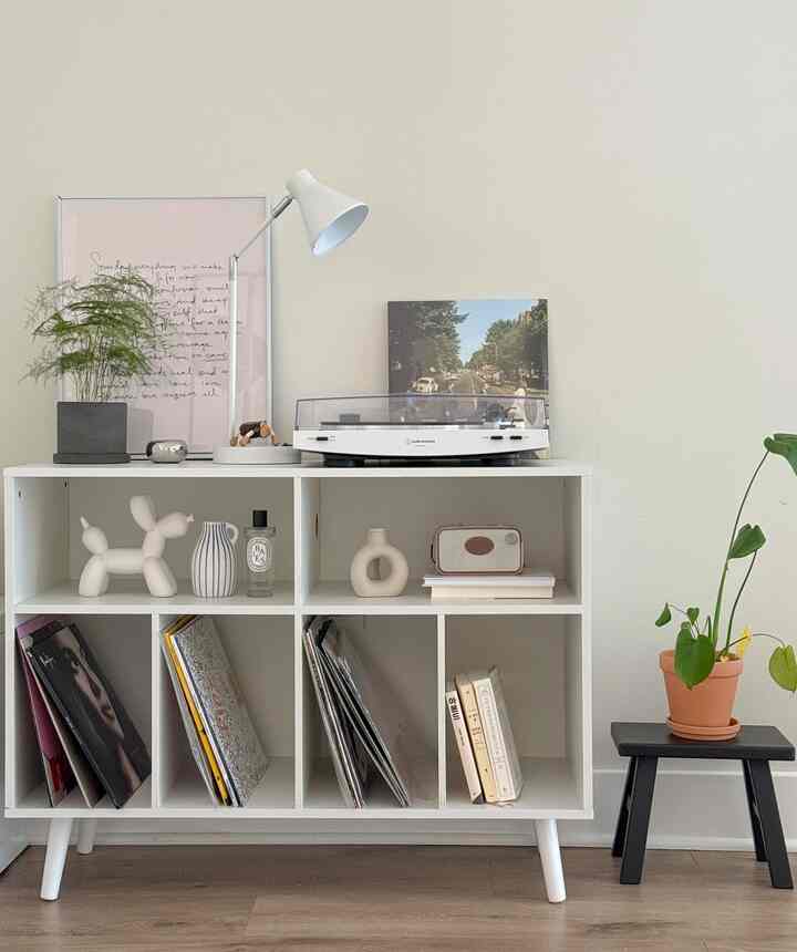 Bright white-toned living space with wood flooring, featuring a white shelf holding a record player and plants, creating a cozy minimalist interior