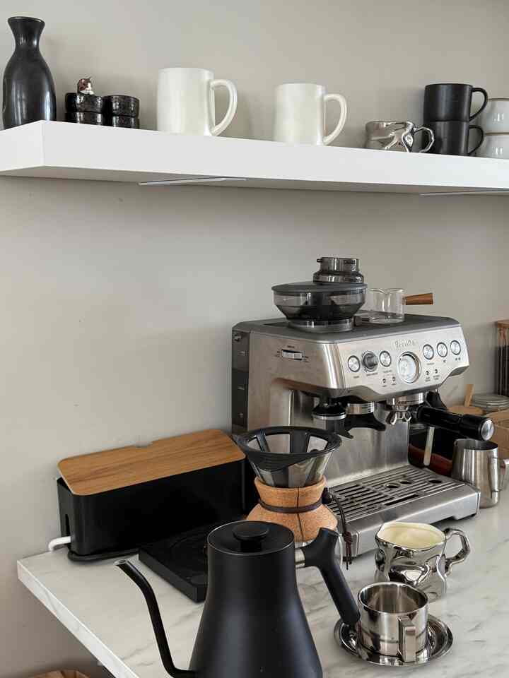 Home cafe space with white walls and shelf, featuring stainless steel coffee machine and various mugs in a clean atmosphere