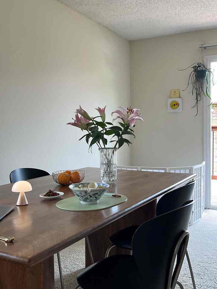 Natural wood tone and white background dining room featuring a table with a vase of flowers and a table lamp in a peaceful setting