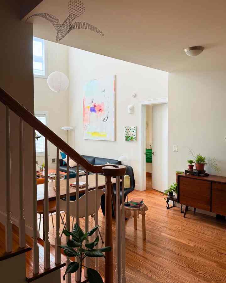 A living room with white walls and wood-tone flooring, featuring a modern dining table, armchairs, and various plants