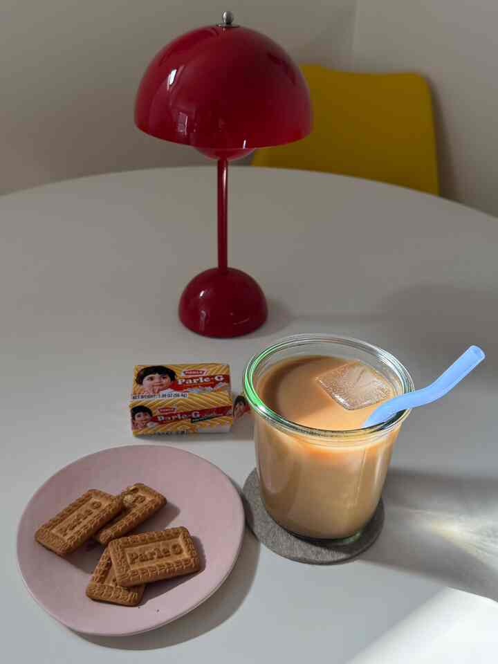 Modern home cafe space with a white dining table featuring a red table lamp and yellow dining chair, relaxing atmosphere with iced chai latte and cookies