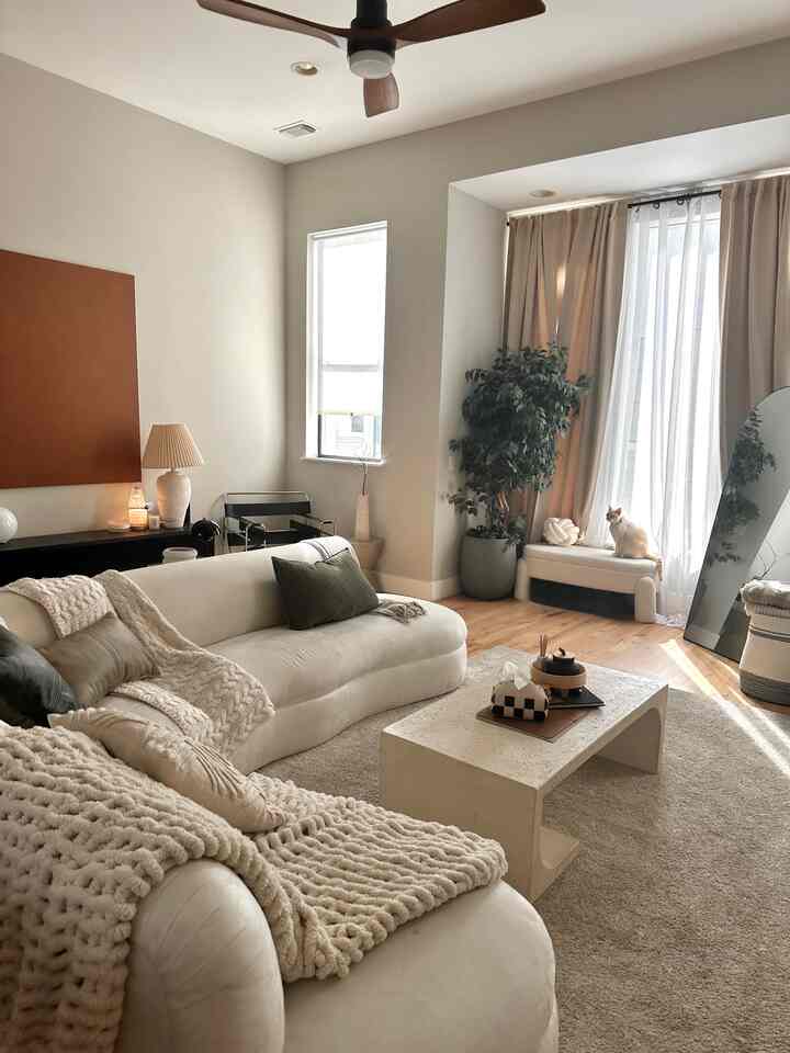 Ivory and beige toned living room featuring a cream sofa, coffee table, large window curtains, and a cat by the window creating a cozy atmosphere