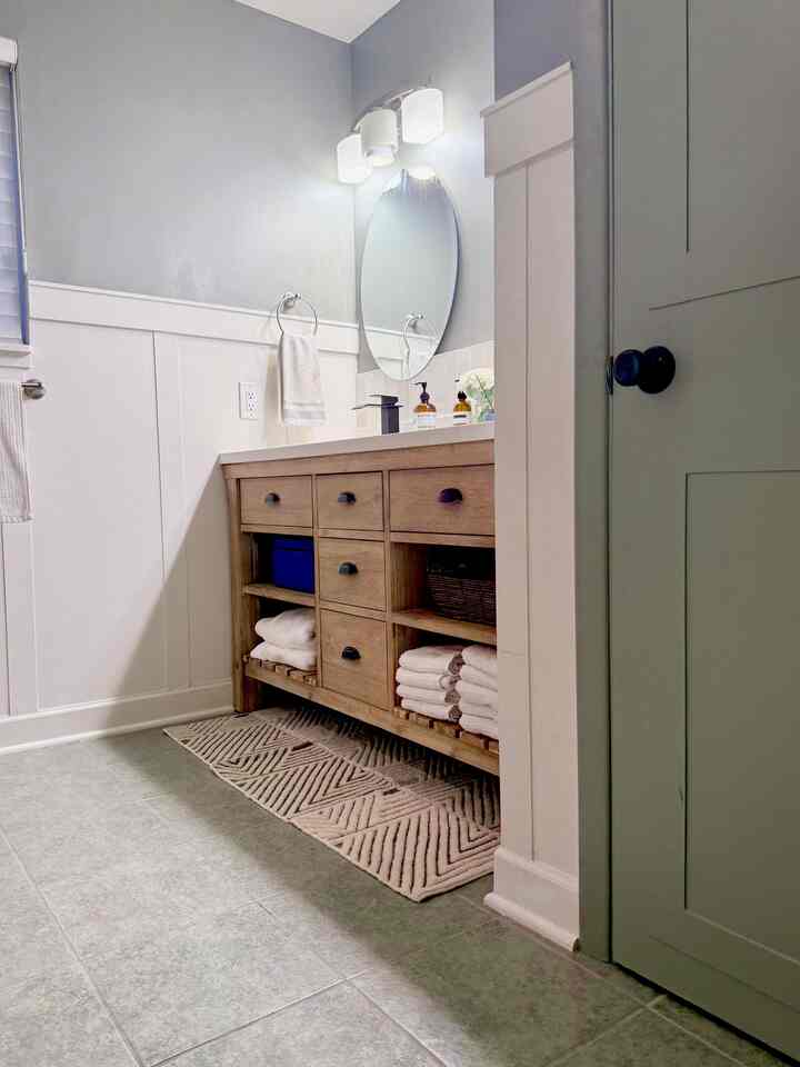 Light gray and green toned bathroom featuring natural wood vanity, white walls, wall sconces, and a bath mat creating a cozy atmosphere
