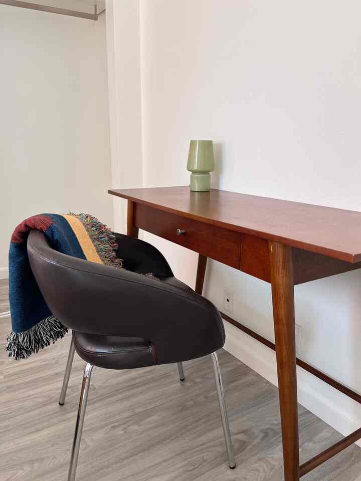 A simple and modern workspace with white walls, brown wood desk, dark brown leather armchair, and a colorful throw blanket