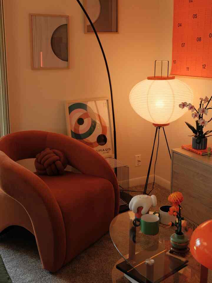 Warm beige-toned living room corner featuring a burnt orange armchair, round tripod floor lamp, and transparent glass coffee table creating cozy atmosphere