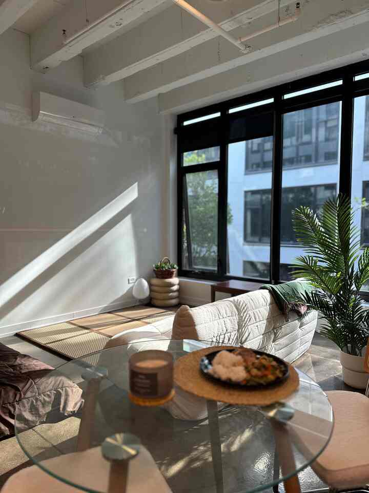 White walls and light brown wood tones define a Japandi-style studio apartment with a sofa, round dining table, and plants in natural spring light