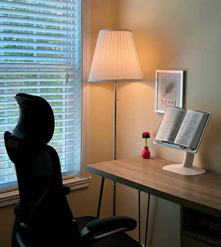 Beige-toned bedroom featuring a simple home office setup with desk, office chair, floor lamp, and a book holder for reading