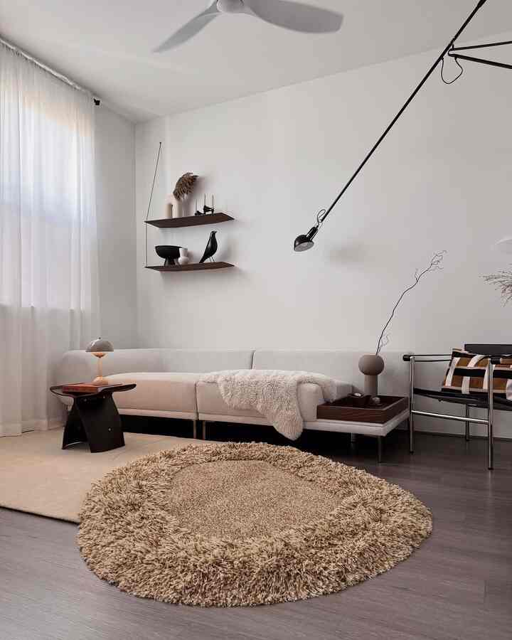 Neutral-toned living room featuring a curved lounge sofa, round shaggy rug, wall shelves, and an adjustable wall lamp in a minimal, calm atmosphere