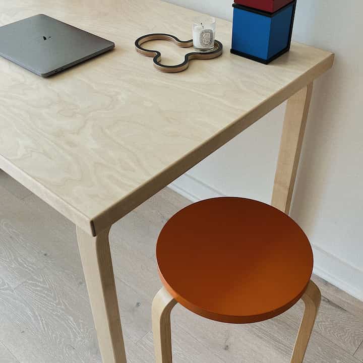 Natural wood tone desk with an orange stool in a minimalist and tidy home office space