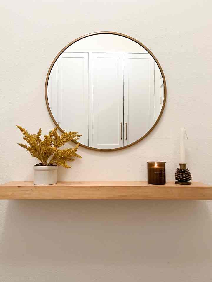White-walled entrance featuring a wood tone floating shelf with a round mirror, yellow dried flowers, and candle creating simple wall decor