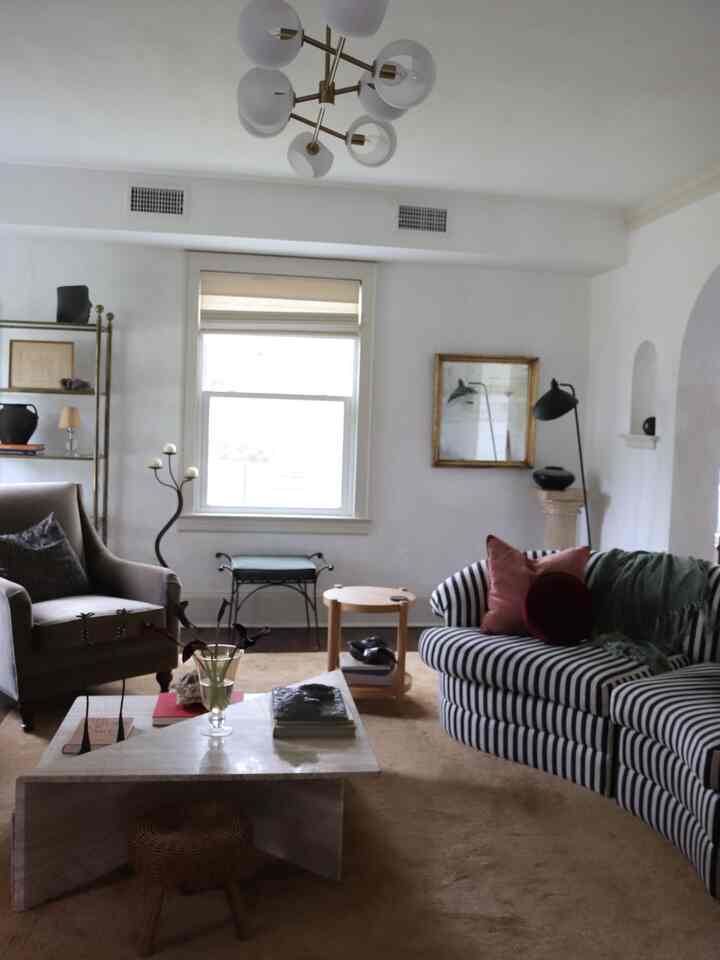 Bright living room with white walls and beige carpet, featuring striped curved sofa and vintage armchair with warm ambiance