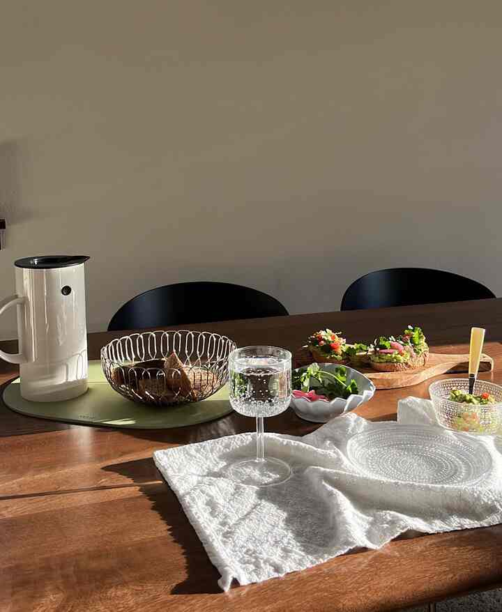 Natural wood tone dining table with white placemat, clear glass, and dining chairs in a simple and cozy dining room space