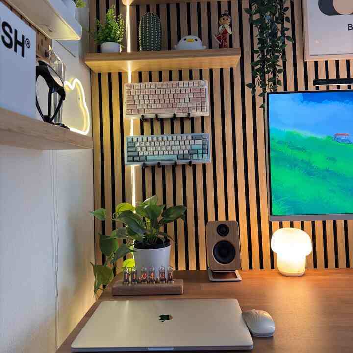 Wood-toned vertical slat wall and white side wall compose a compact home office featuring two wall-mounted keyboards, desk monitor, lighting, and plant creating a cozy atmosphere
