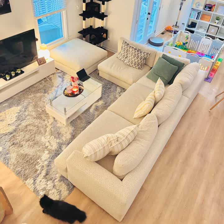 A white and natural toned living room featuring a large beige sectional sofa, kids' play area, and a cat tower creating a cozy atmosphere