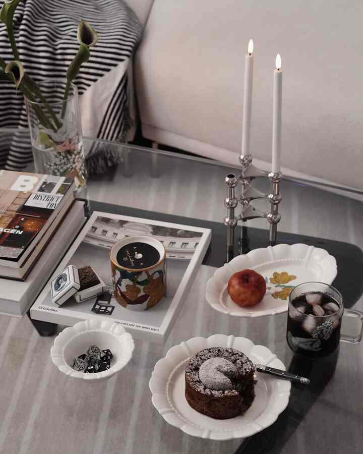 A modern living room with white and transparent glass coffee table, featuring desserts, candles, and a vase