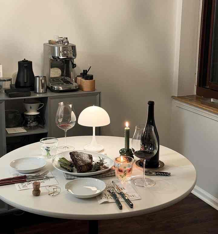 White and wood-toned compact dining room featuring a round dining table with wine glasses and steak, creating a cozy home cafe atmosphere
