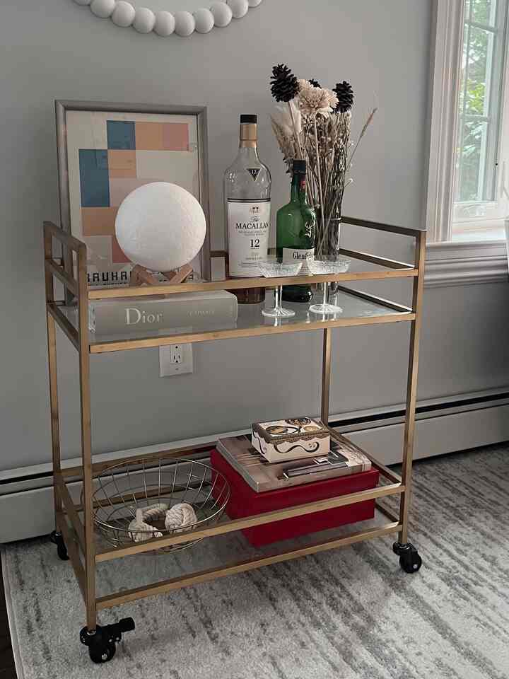Gold tone bar cart in an interior space featuring whiskey bottles, glassware, and a vase with dried flowers, creating a stylish home bar atmosphere