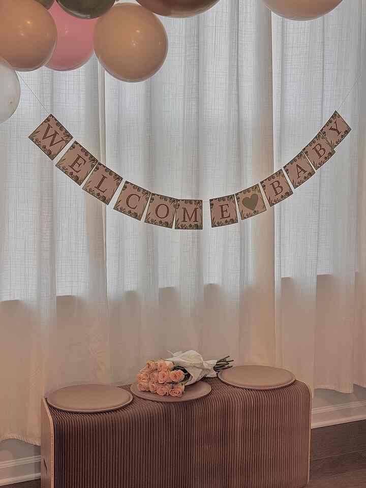Warm welcome baby party space with brown bench holding pink flower bouquet, pastel balloons, and white curtain background