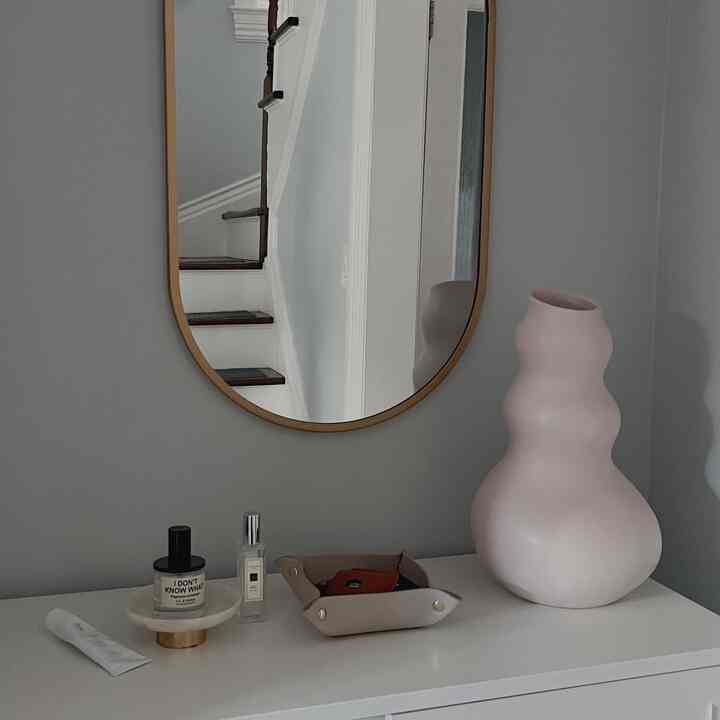Gray-walled space with a white cabinet featuring an oval mirror, a uniquely shaped vase, and a valet tray, creating a calm entrance area