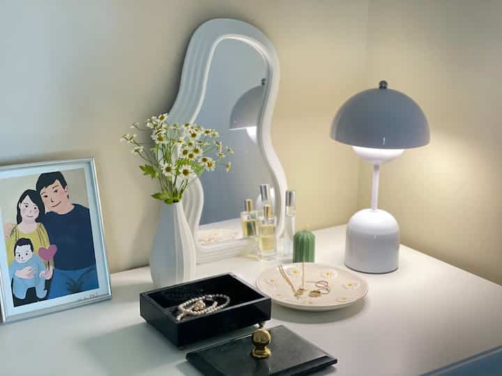 A white and beige toned walk-in closet with a curvy mirror, table lamp, picture frame, and decorative items arranged on a cozy surface