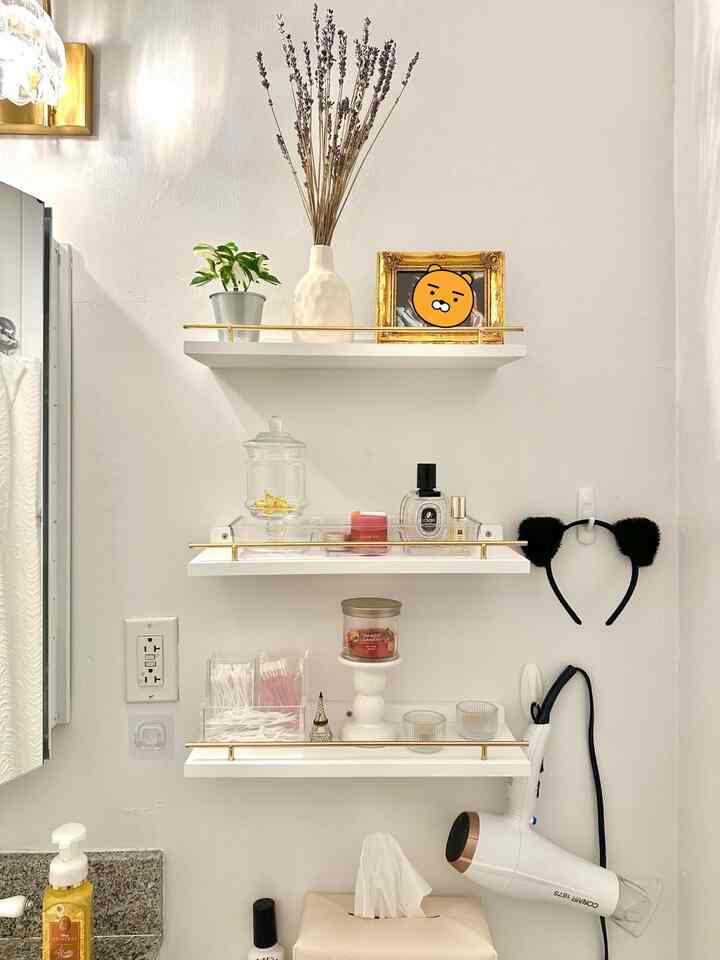White and gold-toned bathroom wall shelves with neatly organized vanity items and beauty tools in a tidy space