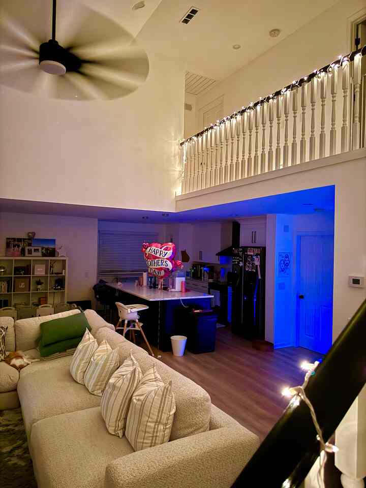 An open-plan living room and kitchen featuring white walls and cabinetry with a beige sofa, cozy string lights, creating a warm ambiance