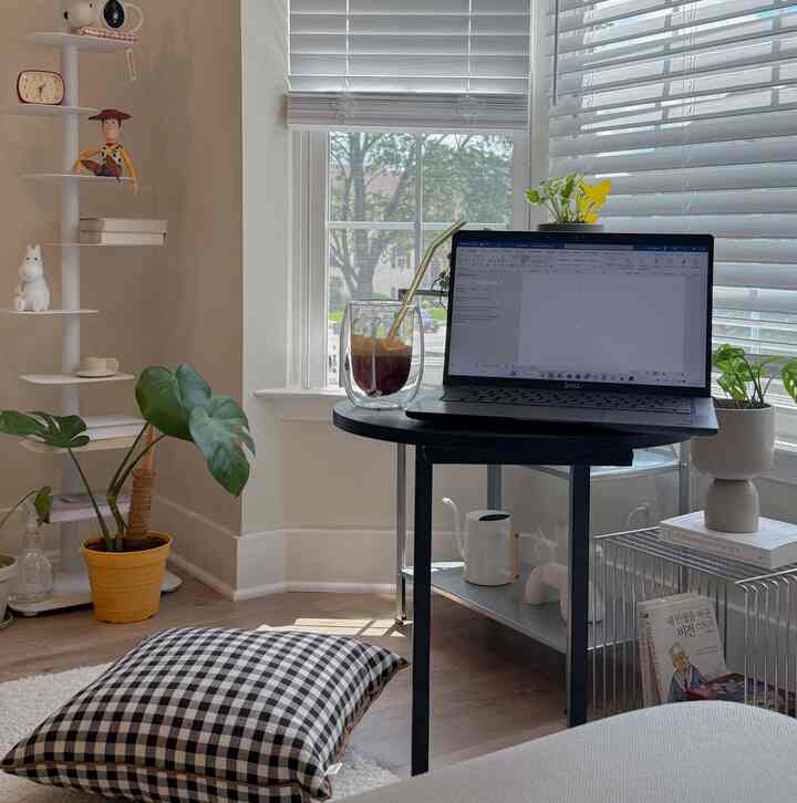 Bright natural-tone home office space featuring a black round desk by the window with plants and a coffee glass, creating a warm atmosphere