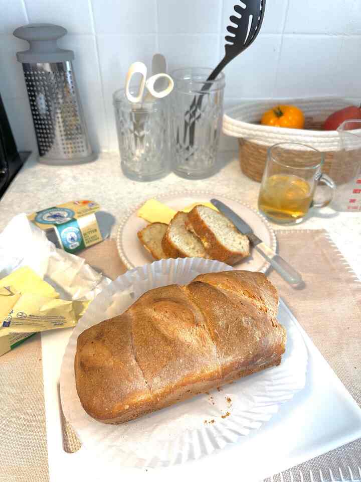 Beige tablecloth covered kitchen countertop with home-baked bread, butter, and a glass of tea against white tiled backsplash