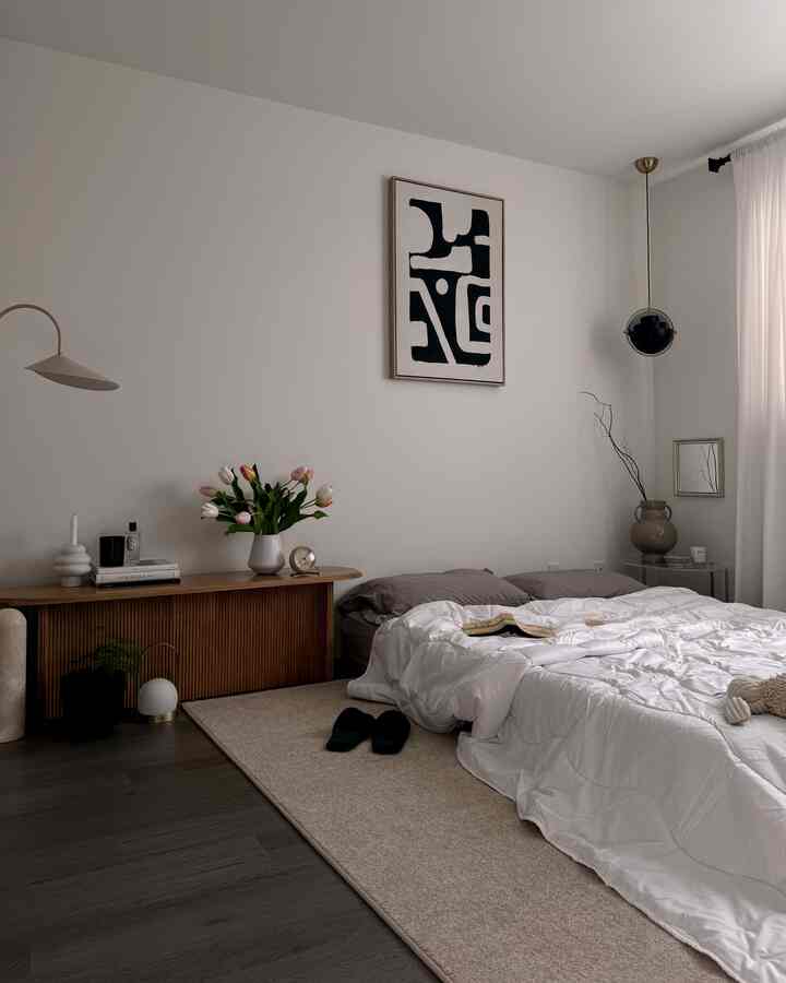 White and brown toned bedroom featuring a simple bed, rug, vase with flowers, and abstract wall art in a Japandi style setting