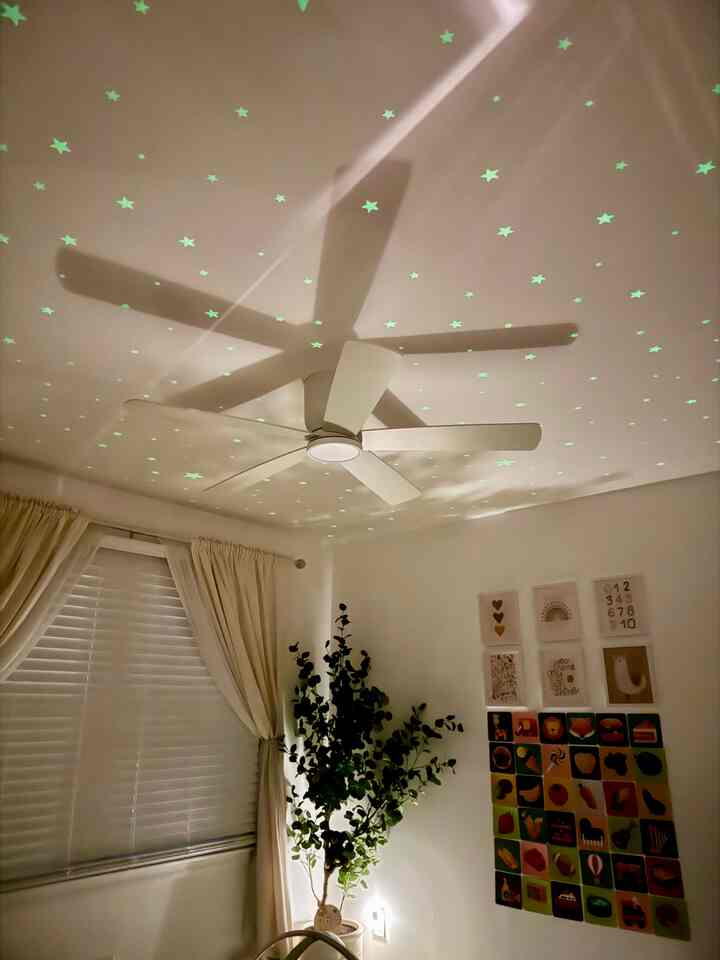 A white and natural tone kids' room with a ceiling fan featuring star lights, beige curtains, and a eucalyptus plant creating a calm atmosphere