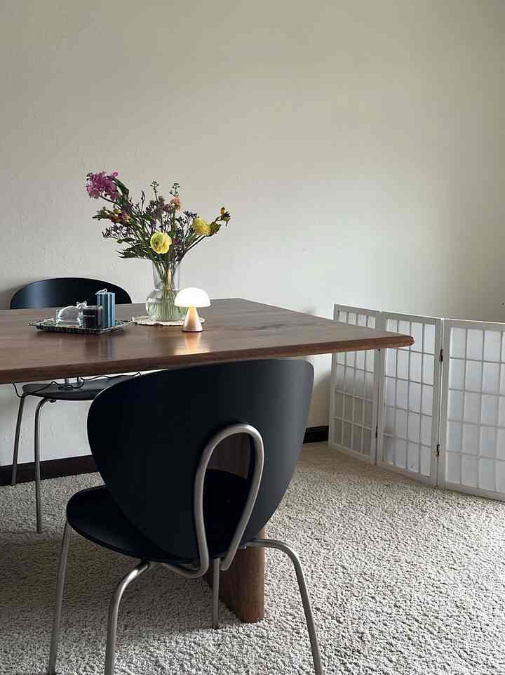 Natural-toned dining room featuring a wooden dining table, black chairs, a vase with flowers, and a small table lamp, creating a cozy atmosphere