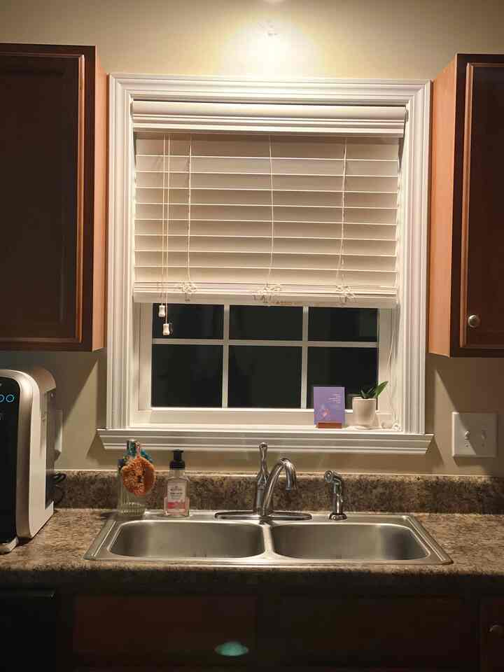 Beige-toned kitchen with wood tone cabinets, featuring a central double sink and chrome kitchen faucet under a white blind-covered window