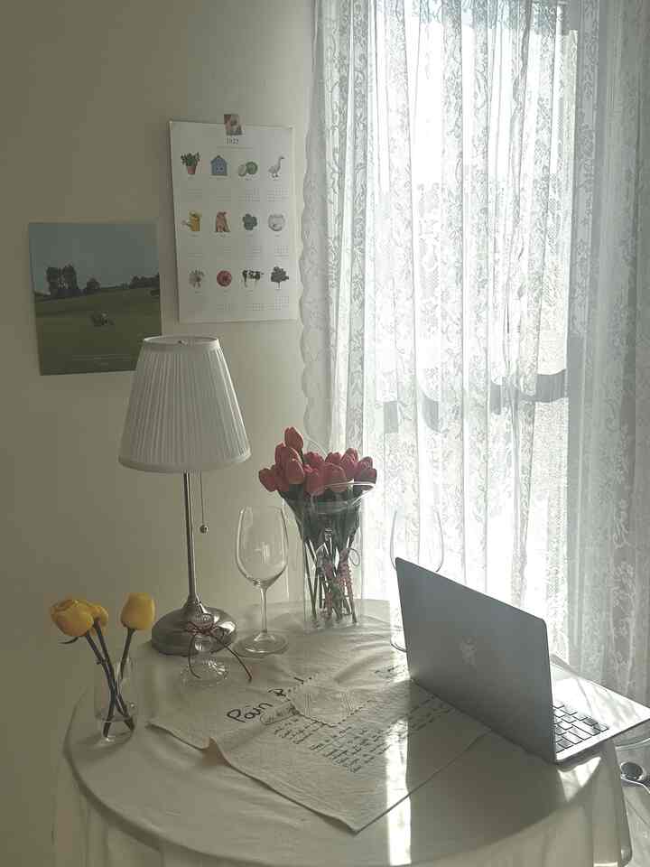 White and beige toned home office space featuring table lamp, vase with flowers, and laptop, creating a simple and cozy atmosphere