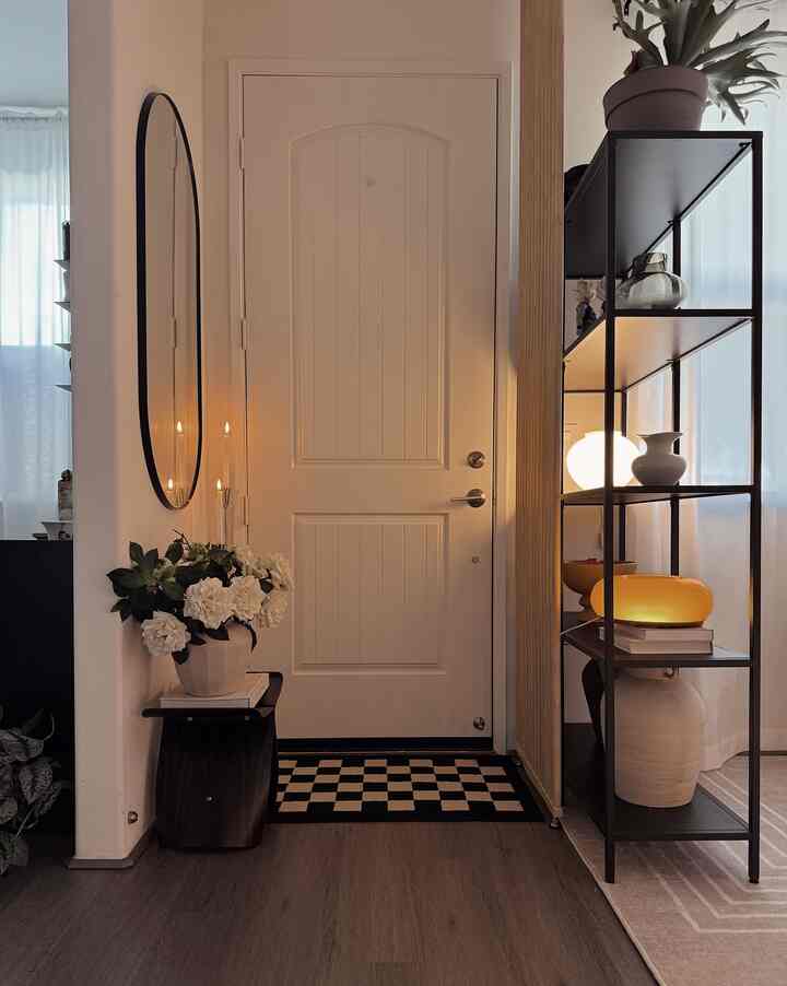 A small entrance in white and black tones featuring a stool with vase and shelf lighting for a chic atmosphere