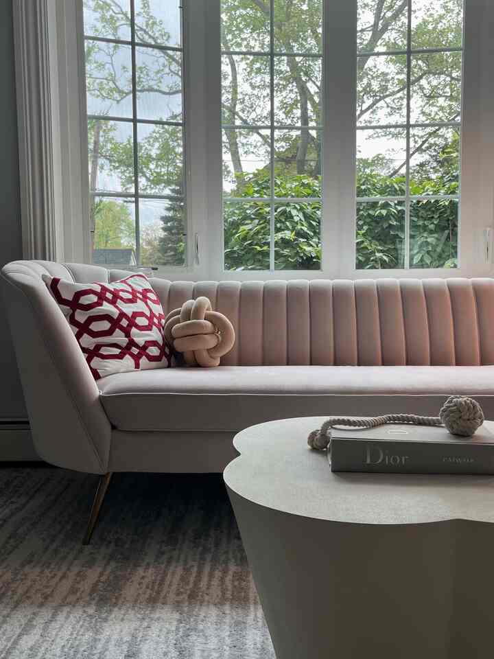 A simple and cozy living room featuring a pink sofa and gray rug with large windows showcasing green plants outside