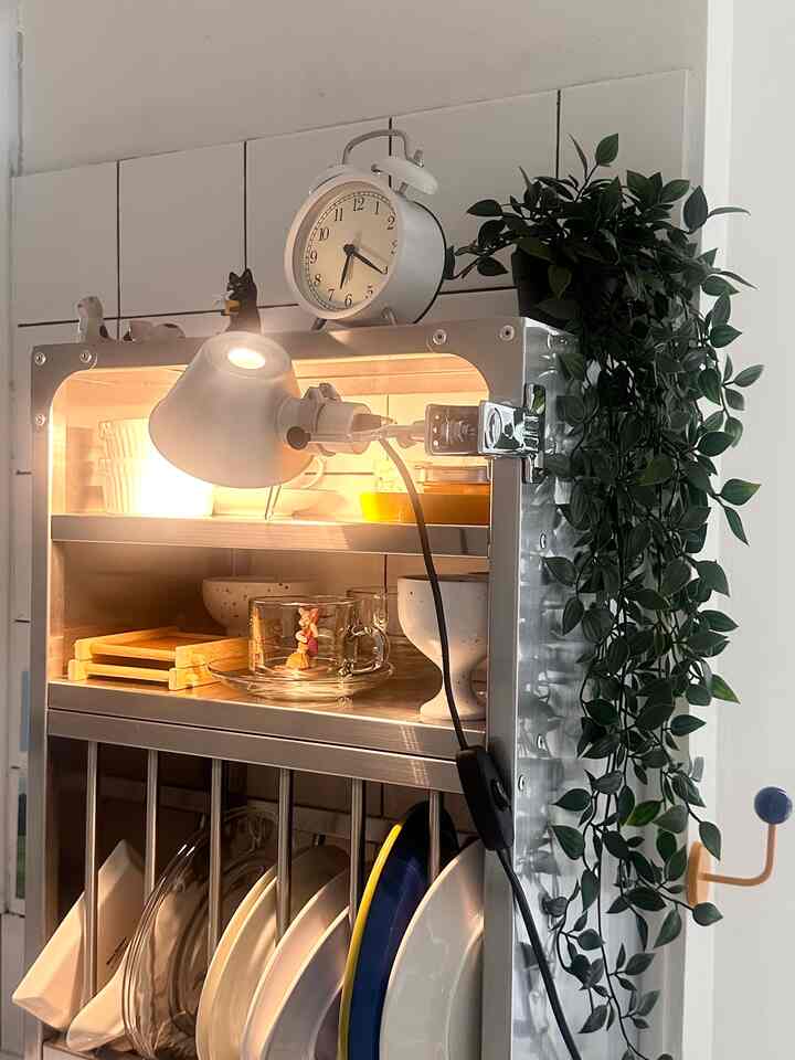 Minimal kitchen space with white and metal tones, neatly arranged plates and bowls, decorated with a plant and warm lighting