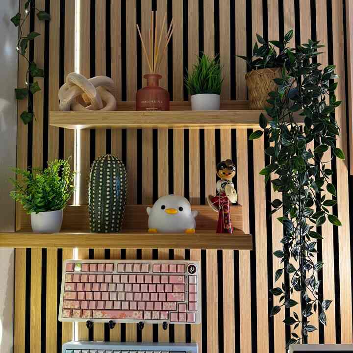 Minimal home office space featuring light wood tone slatted wall shelves with plants, diffuser, objet, and a pink keyboard