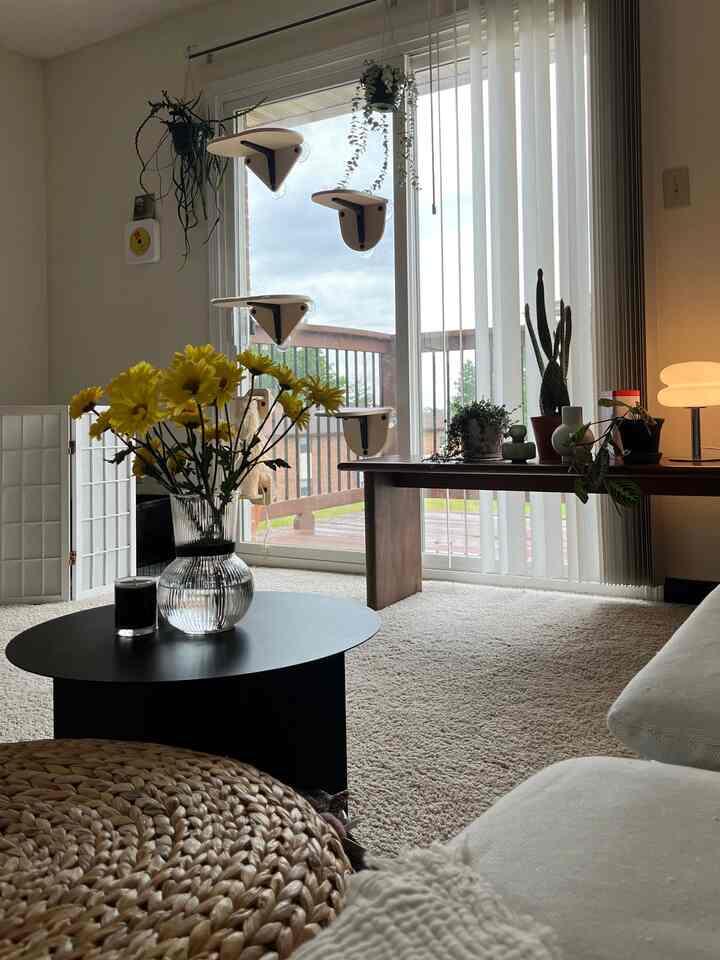 Beige-carpeted living room with white sofa, black coffee table in center featuring yellow flowers and candle, creating a cozy atmosphere