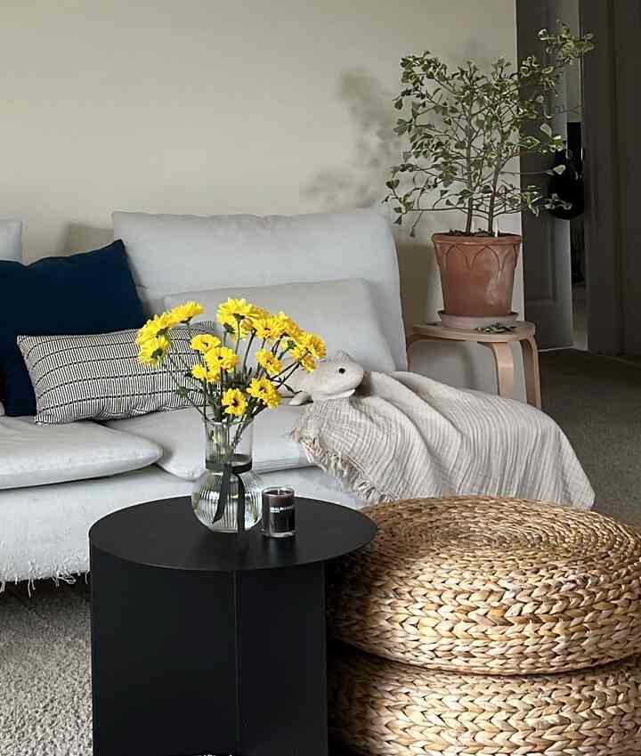 White and natural tone living room featuring a white sofa, yellow flowers in a glass vase, woven stools, and a wooden side table creating a cozy atmosphere