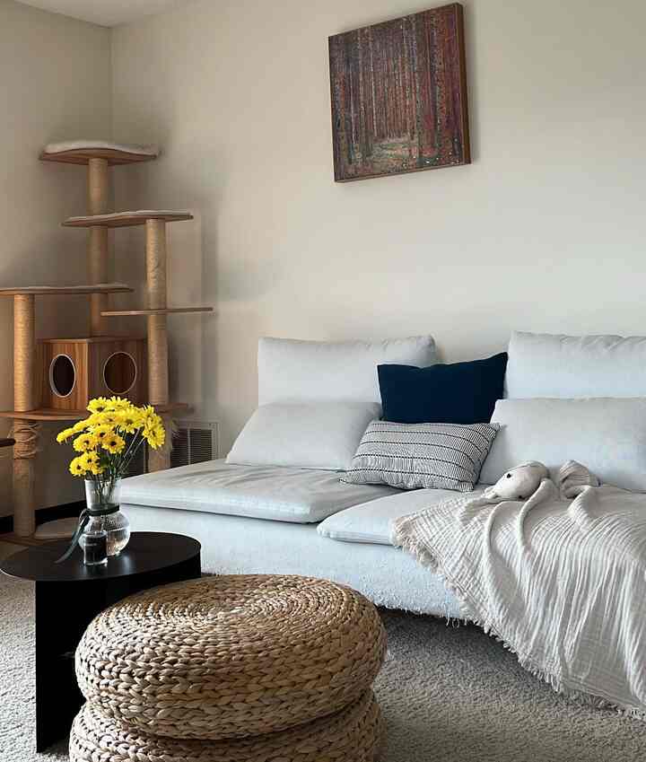 Beige-toned living room featuring white sectional sofa with navy cushion, cat tower, and natural woven ottoman in a simple setting